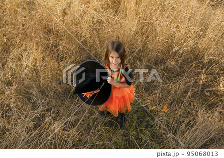 A girl dressed as a witch holds a huge black hat with an orange ribbon and looks up smiling sweetly. Waiting for Halloween Day A girl dressed as a witch holds a huge black hat with an orange ribbon and looks up smiling sweetly. Waiting for Halloween Day 95068013