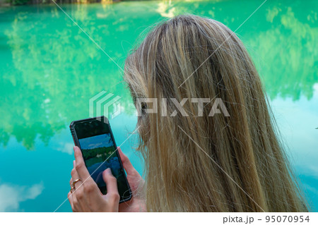 Young girl near lake taking a photo with a smartphone while. Happy attractive girl on the beach in the day, back view 95070954
