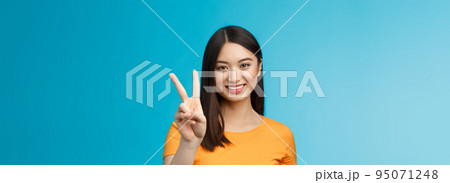 Cheerful friendly asian woman in yellow t-shirt smiling broadly, show number two, twice, win second place, smiling joyfully, make take-away order, book sits, stand blue background 95071248