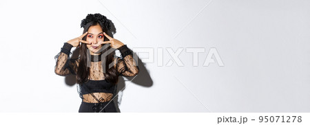 Beautiful asian woman in halloween party outfit looking at upper left corner, making peace gestures over eyes, standing in gothic lace dress with black wreath over white background 95071278
