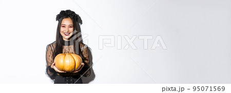 Happy beautiful woman in black lace dress enjoying halloween holiday, smiling at camera and holding pumpkin, standing over white background 95071569