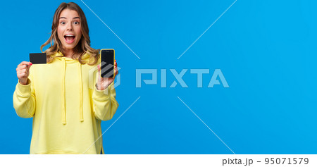 Amused and excited blond girl recommend using banking service, showing smartphone display and credit card and looking camera fascinated as receive bonus to bank account, blue background 95071579