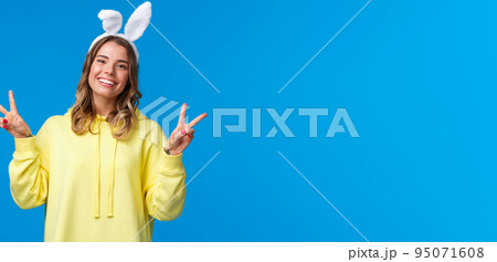 Holidays, traditions and celebration concept. Kawaii young blond girl in rabbit ears showing peace gesture and smiling, having fun, enjoying party, standing blue background 95071608