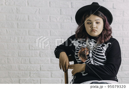Young Asian teen in costume witch sitting on a chair in front of a white brick wall, background of Halloween. Young Asian teen in costume witch sitting on a chair in front of a white brick wall, background of Halloween. 95079320