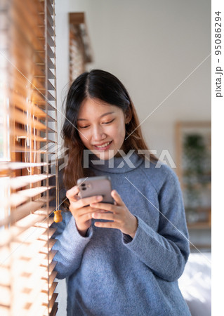 Cheerful young asian woman using mobile phone near window at home. 95086294