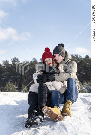 Young guy and a girl sit and hug on the snow against the background of the forest and the blue sky. Vertical frame Young guy and a girl sit and hug on the snow against the background of the forest and the blue sky. Vertical frame 95086690