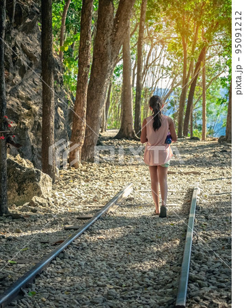 Back view of young girl with water bottle walking alone in forest nature path walk on trail woods background at Hellfire Pass in Kanchanaburi Thailand. Happy girl relaxing on active outdoor activity. 95091212