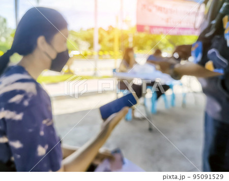 Blurred image of young woman wear surgical face mask use smart phone while waiting in a queue to take first dose of the COVILO (Sinopharm) Coronavirus vaccine during vaccination drive to all adults. 95091529