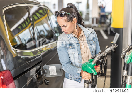 Woman holding gas and diesel pistols in the service station looks at the open gas tank of her car Woman holding gas and diesel pistols in the service station looks at the open gas tank of her car 95091809