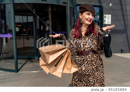 A happy shopaholic girl keeps her bags near the shopping center. A woman near the store is happy with her purchases, holding bags. Dressed in a leopard print dress. Consumer concept. 95105894