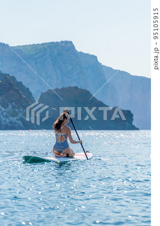 Vertical photo. Sporty girl on a glanders surfboard in the sea on a sunny summer day. In a striped swimsuit, it is in the bar. Summer activities by Stortom by the sea Vertical photo. Sporty girl on a glanders surfboard in the sea on a sunny summer day. In a striped swimsuit, it is in the bar. Summer activities by Stortom by the sea 95105915
