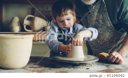 Curious little boy is learning pottery from his experienced grandfather in small home studio. Child is forming clay to make pot on potter's wheel, his granddad is helping him. Curious little boy is learning pottery from his experienced grandfather in small home studio. Child is forming clay to make pot on potter's wheel, his granddad is helping him. 95106092