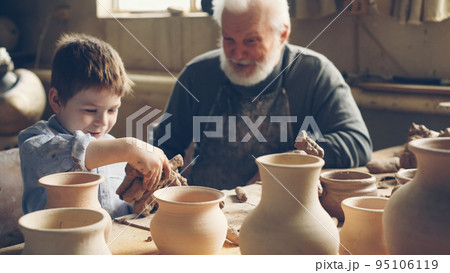 Cute little boy is playing with clay sitting with his grandparent at table in home pottery studio. Grandfather is talking to curious child, boy is enjoying new hobby. Cute little boy is playing with clay sitting with his grandparent at table in home pottery studio. Grandfather is talking to curious child, boy is enjoying new hobby. 95106119