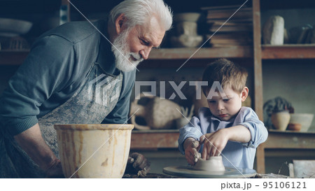 Concentrated young boy is molding clay into ceramic pot on spinning throwing wheel and his loving experienced grandfather is talking to him. Pottery and family tradition concept. Concentrated young boy is molding clay into ceramic pot on spinning throwing wheel and his loving experienced grandfather is talking to him. Pottery and family tradition concept. 95106121