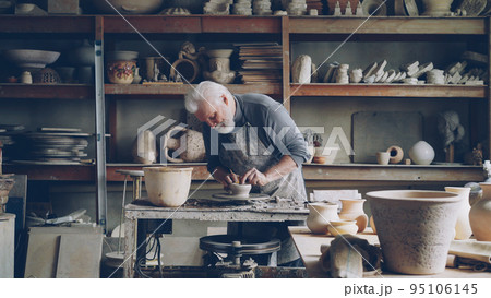 Experienced male ceramist is creating utensils from clay on potter's wheel in workshop. Producing eathenware, handmade utensils and professional pottery concept. 95106145