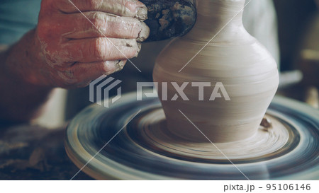 Elderly bearded craftsman is creating ceramic jar on potter's wheel using professional tools. Workplace with handmade pots, beautiful vases and figures in background. 95106146