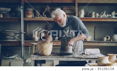 Hardworking grey-haired man is working with clay on potter's wheel in workshop, shaping piece of loam. Beautiful ceramic utensils, handmade pots and vases on shelves are visible. Hardworking grey-haired man is working with clay on potter's wheel in workshop, shaping piece of loam. Beautiful ceramic utensils, handmade pots and vases on shelves are visible. 95106158