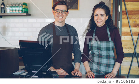 Portrait of two young good-looking waiters standing at cashier's desk in modern coffee-house and smiling. Successful business, happy people and food service concept. 95106182