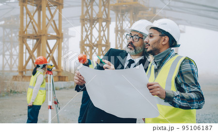 Architect man in suit is talking to engineer in building area while surveyors are working with modern equipment in background. People and business concept. 95106187