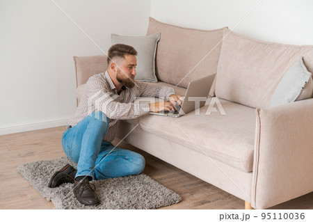 Young bearded man smiles while reading laptop screen while relaxing Young bearded man smiles while reading laptop screen while relaxing 95110036