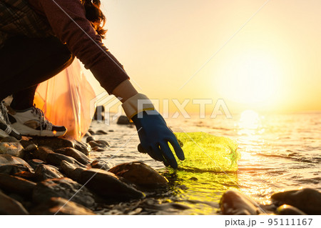 Coastal cleanup and garbage collection for recycling. A woman volunteer collects plastic bottle by the sea or river, closeup of hand. Low angle view. World environment day and Earth day concept 95111167