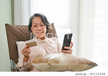 Senior Asian female using mobile banking application while relaxing in her living room. Senior Asian female using mobile banking application while relaxing in her living room. 95115346