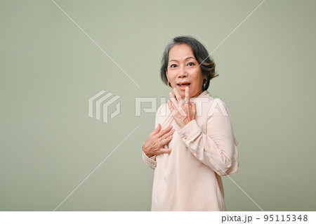 Surprised and excited 60s senior Asian woman standing against isolated green background Surprised and excited 60s senior Asian woman standing against isolated green background 95115348