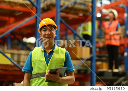 Portrait of male warehouse worker wearing safety hardhat and vest holding digital tablet standing in large retail warehouse 95115950