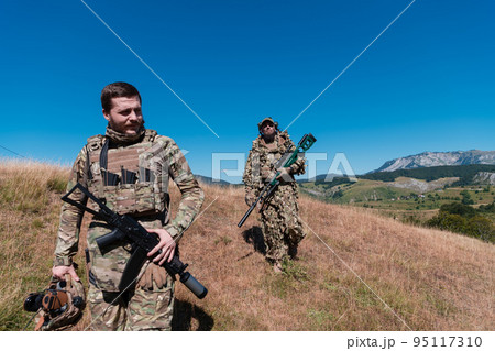 A sniper team squad of soldiers is going undercover. Sniper assistant and team leader walking and aiming in nature with yellow grass and blue sky. Tactical camouflage uniform. 95117310