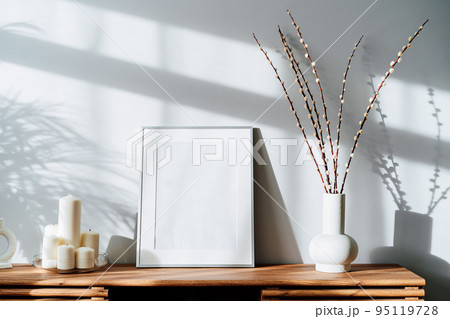 Modern minimalist style interior with white poster mockup, candles and blooming branches of the pussy willow in vase on wooden console under sunlight and shadows on a white gray wall. Selective focus 95119728