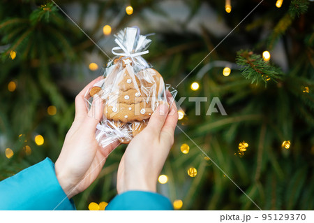 A woman holds in her hands gingerbread in the form of a Christmas tree on the background of a green Christmas tree outdoors. The concept of winter holidays, sweets and coziness. 95129370