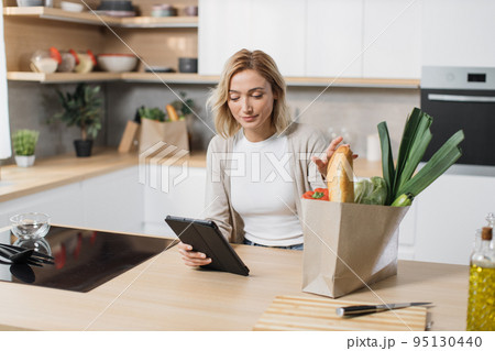Woman cooking healthy food looking on paper bag with vegetables while reading recipe on tablet Woman cooking healthy food looking on paper bag with vegetables while reading recipe on tablet 95130440