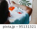 Cute child siting at desk and drawing rowanberries on album sheet with dry rowan leaves 95131321