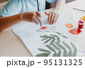 Cute child siting at desk and drawing rowanberries on album sheet with dry rowan leaves 95131325