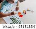 Cute child siting at desk and drawing rowanberries on album sheet with dry rowan leaves 95131334