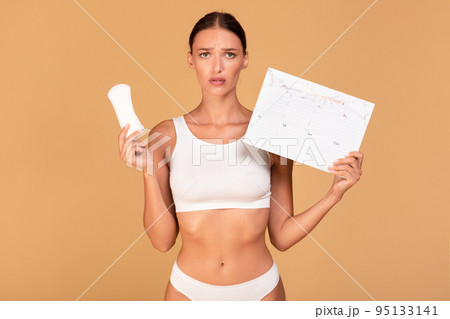Period and female hygiene protection. Upset woman in white underwear holding menstrual pad and paper calendar Period and female hygiene protection. Upset woman in white underwear holding menstrual pad and paper calendar 95133141