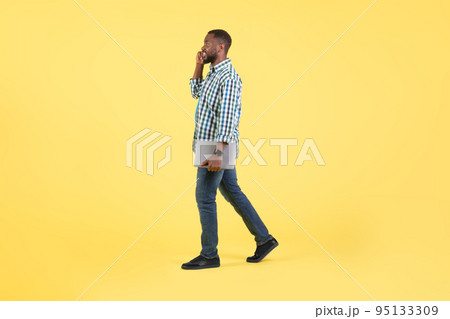 African Male Talking On Phone Holding Laptop Walking, Yellow Background 95133309