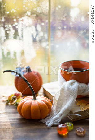 An orange mug in a scarf with hot tea, pumpkins, yellow dry maple leaves, a book on the windowsill, raindrops on the window - autumn mood An orange mug in a scarf with hot tea, pumpkins, yellow dry maple leaves, a book on the windowsill, raindrops on the window - autumn mood 95141137
