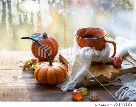 An orange mug in a scarf with hot tea, pumpkins, yellow dry maple leaves, a book on the windowsill, raindrops on the window - autumn mood An orange mug in a scarf with hot tea, pumpkins, yellow dry maple leaves, a book on the windowsill, raindrops on the window - autumn mood 95141138