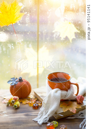 An orange mug in a scarf with hot tea, pumpkins, yellow dry maple leaves, a book on the windowsill, raindrops on the window - autumn mood An orange mug in a scarf with hot tea, pumpkins, yellow dry maple leaves, a book on the windowsill, raindrops on the window - autumn mood 95141141