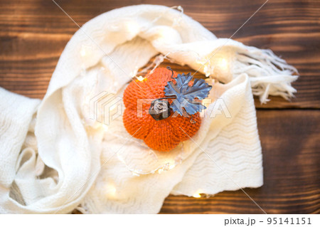Orange pumpkin wrapped in a warm white plaid with lights of garlands on a wooden table with copyspace. Autumn mood, halloween, harvest, thanksgiving, home decor and cozy. Selective focus 95141151