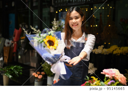 Smiling young female florist holding a bouquet of beautiful colorful flowers standing front of her floral shop 95145665