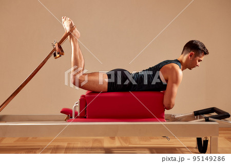 Fit man doing a lunge stretch yoga pilates exercise to strengthen and tone his muscles using a reformer in a gym Fit man doing a lunge stretch yoga pilates exercise to strengthen and tone his muscles using a reformer in a gym 95149286