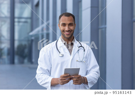Portrait of happy and smiling hispanic doctor, man smiling and looking at camera, doctor using tablet computer for online consultation, outside of modern clinic. 95163143
