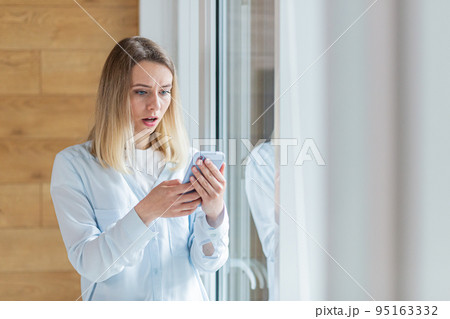 young woman saw bad news on smartphone, standing by the window in the office or at home indoors. The female anxiously holds a mobile phone in her hands, reads the news of the day 95163332