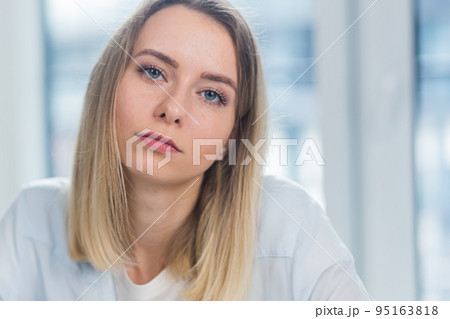 Close-up portrait of a young adult blonde business woman looking at the camera indoors at office or home on a blue window background. Face Confident female Manager in Formal Wear indoors Close-up portrait of a young adult blonde business woman looking at the camera indoors at office or home on a blue window background. Face Confident female Manager in Formal Wear indoors 95163818