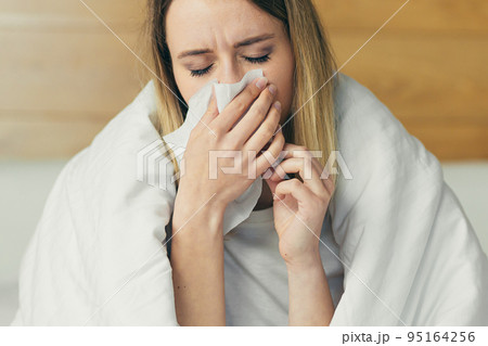 Young woman sitting on the bed at home covered with a blanket, sick, holding his head, has a runny nose, sneezes, allergies, feels bad 95164256