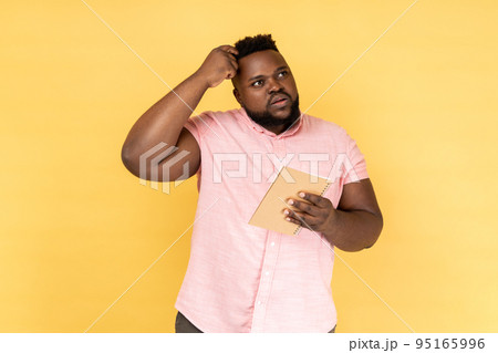 Portrait of thoughtful man wearing pink shirt thinking over smart idea, holding pencil and notebook to write plans, to-do list. Indoor studio shot isolated on yellow background. 95165996