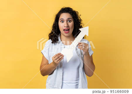 Portrait of amazed excited woman with dark wavy hair standing holding white arrow pointing aside, looking at camera with open mouth. Indoor studio shot isolated on yellow background. 95166009