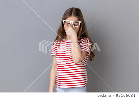 Portrait of sick tired little girl wearing striped T-shirt standing ribbing her eyes, takes off glasses, felling eyes hurt and headache. Indoor studio shot isolated on gray background. Portrait of sick tired little girl wearing striped T-shirt standing ribbing her eyes, takes off glasses, felling eyes hurt and headache. Indoor studio shot isolated on gray background. 95166031